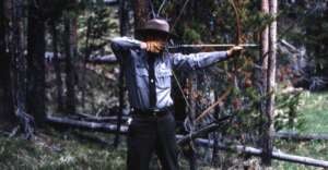A person wearing a ranger uniform and wide-brimmed hat stands in a forest, aiming a bow and arrow, preparing to shoot. Trees and grass are visible in the background.
