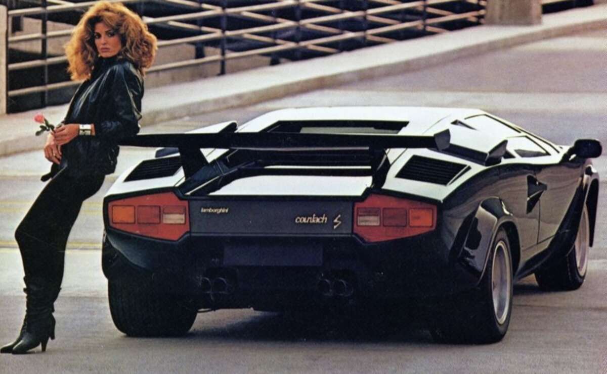 A woman with voluminous hair in a black outfit leans against the rear of a black Lamborghini Countach parked in an urban setting, holding a rose and looking toward the camera.
