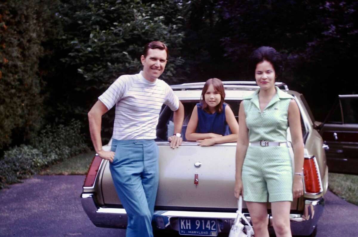 A man, a woman, and a girl pose in front of a beige station wagon parked on a driveway, with trees in the background. The man is smiling, the woman holds a white purse, and the girl leans on the car.