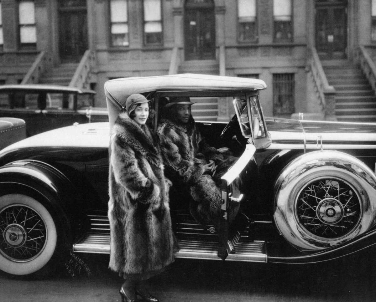 A woman in a fur coat stands next to a man seated in a classic car, both dressed in 1920s fashion, on a city street lined with brownstone buildings.