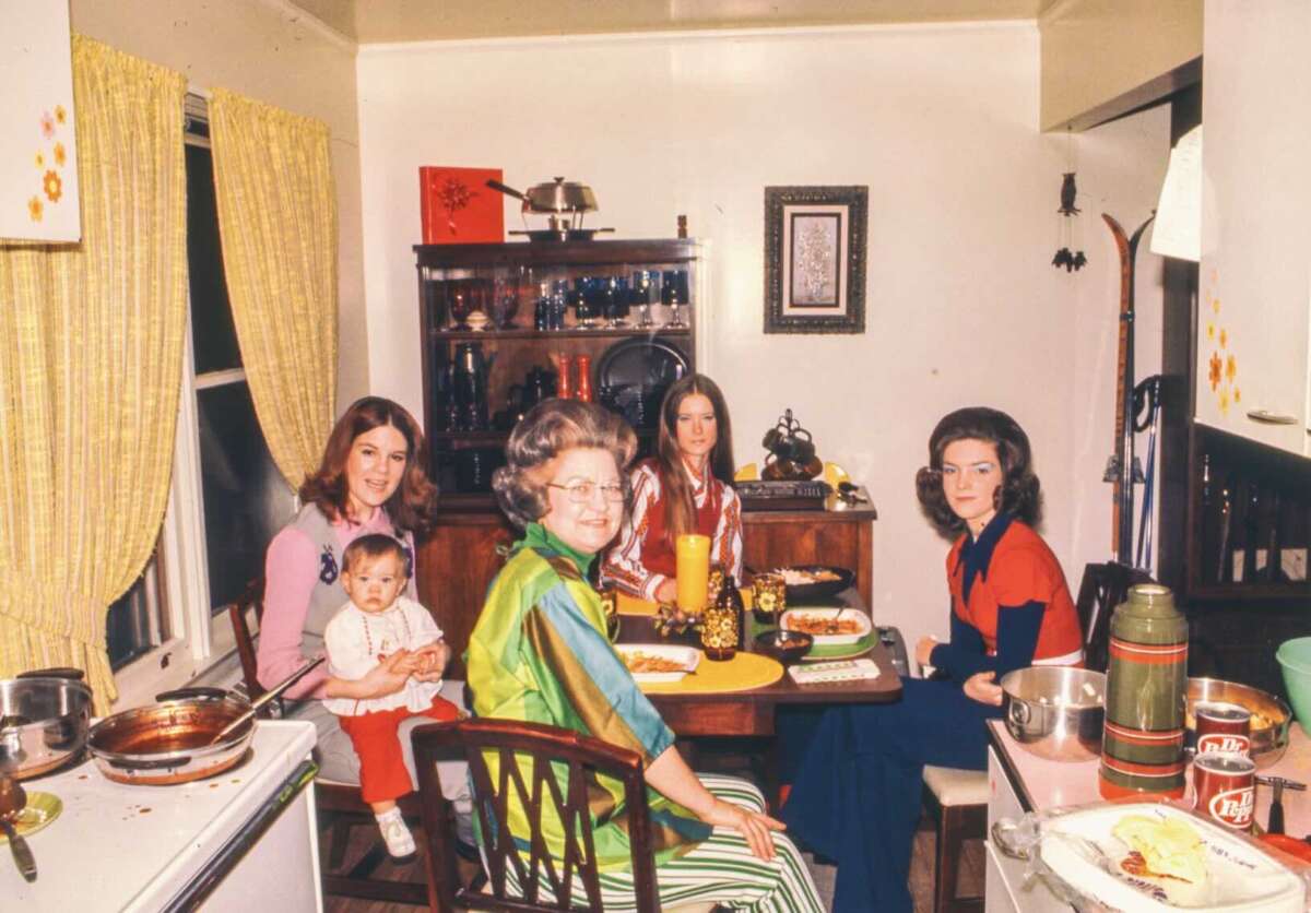 Four women and a baby sit around a table set for a meal in a vintage kitchen with yellow curtains and retro decor. Food, drinks, and dishes are spread out, creating a cozy, homey atmosphere.