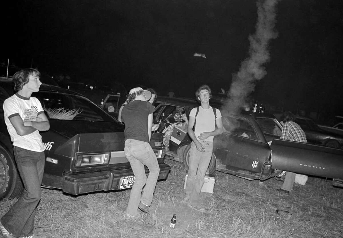 Four people stand near parked cars at night in a grassy area. One person leans on a car, another appears to be dancing or posing, and smoke rises nearby. A beer bottle is on the ground in the foreground.