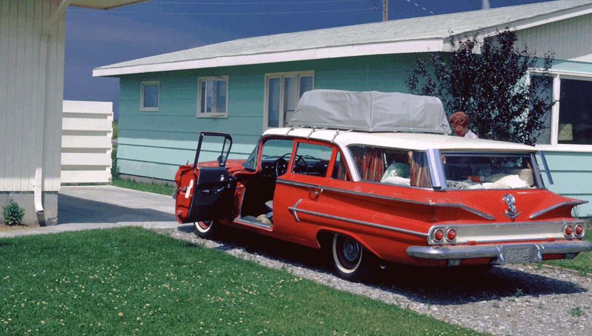 A vintage red station wagon with a roof bag is parked outside a pastel green house. The car doors are open, and a person is loading or unloading items from the back seat on a sunny day.