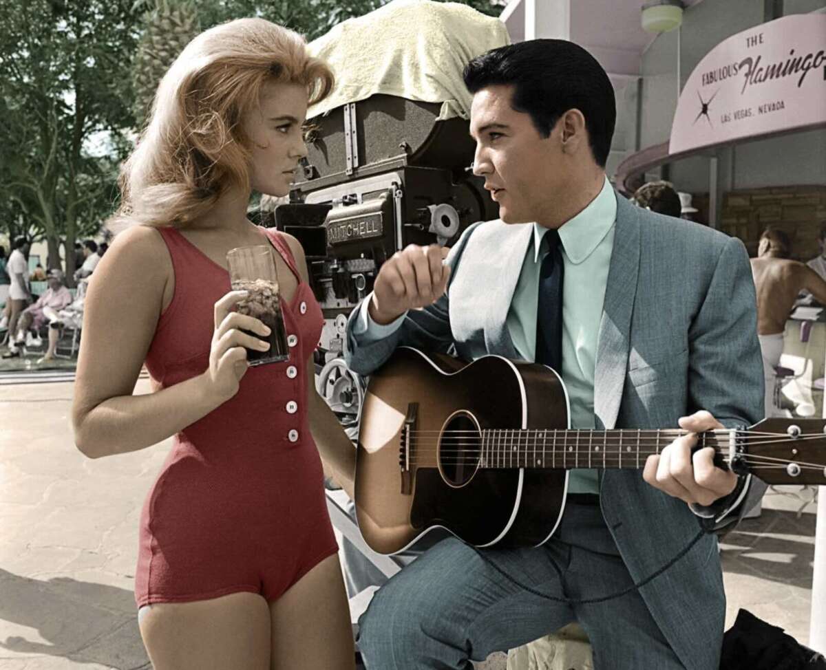 A woman in a red swimsuit holding a drink stands next to a man in a suit playing a guitar. They appear to be conversing outdoors, with a retro camera and people in the background.