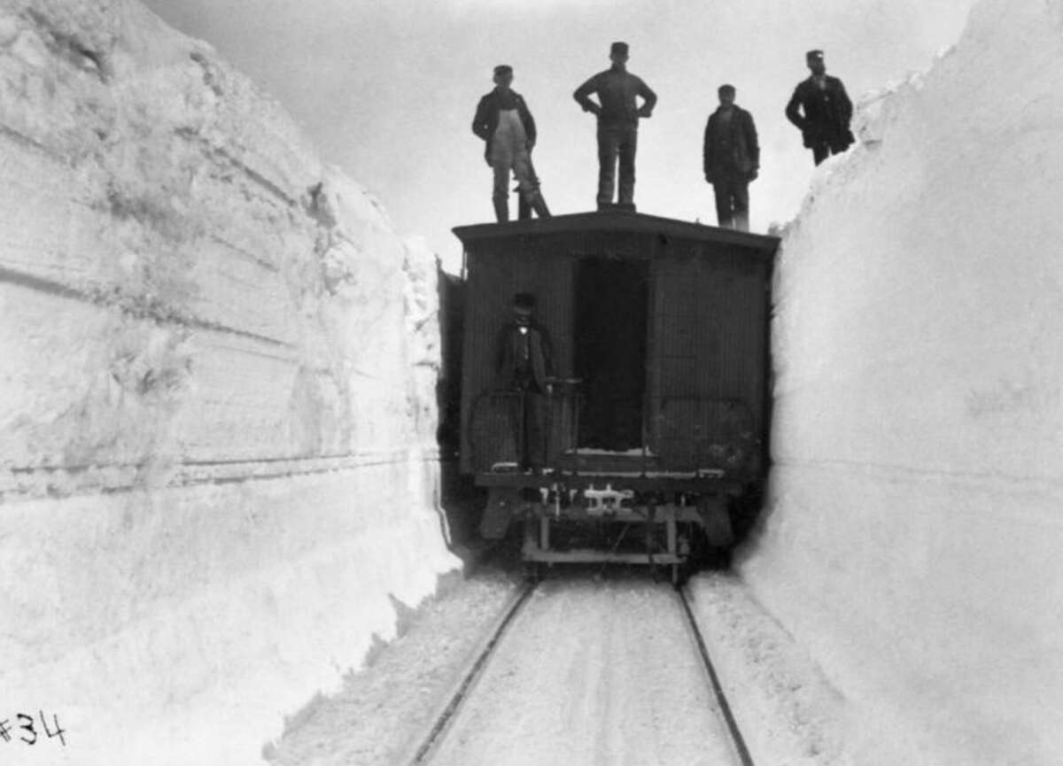 A black-and-white photo shows a train car on tracks surrounded by towering walls of snow, with five people standing on top of the snowbanks and the train car.