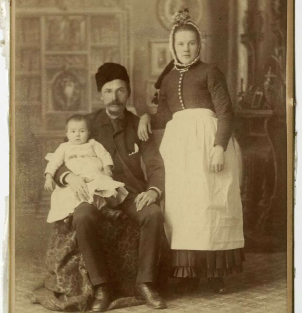 A sepia-toned vintage photograph of a man seated with a child on his lap and a woman standing beside him. They wear traditional 19th-century European clothing, and the background is ornately decorated.