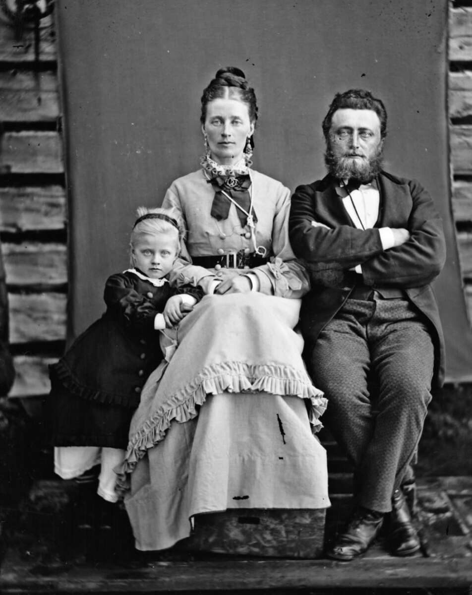 A black-and-white vintage portrait of a family with a woman seated in the center, a man with folded arms sitting to her right, and a young girl leaning against her on the left, all dressed in 19th-century clothing.