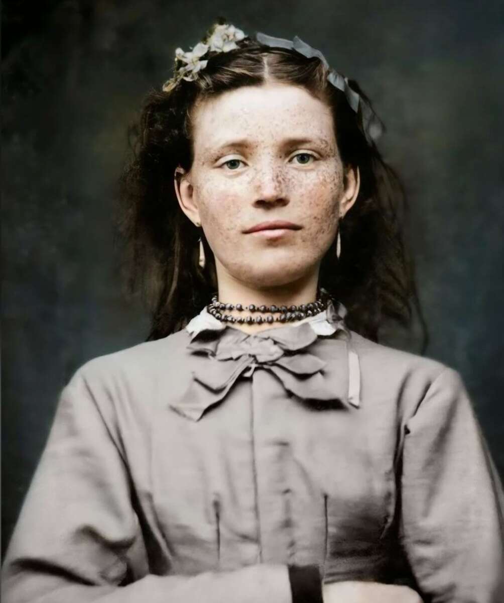 A young woman with fair skin, freckles, and wavy brown hair wears a gray dress with a high collar and a bow, a beaded necklace, and flower hairpins, looking directly at the camera against a dark background.