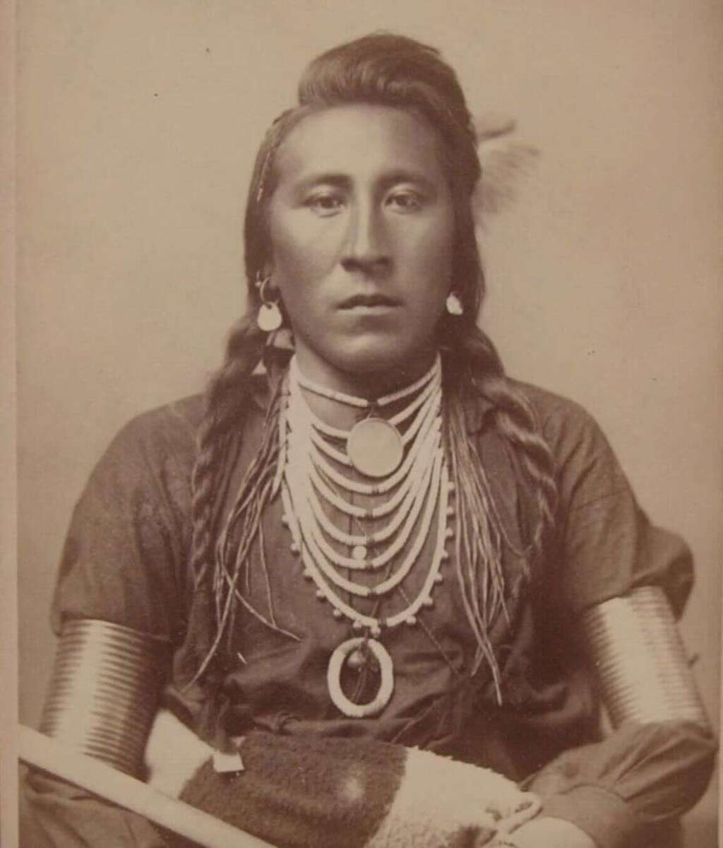 A Native American man with long braids, wearing traditional jewelry, including beaded necklaces and large circular ornaments. He is dressed in a decorated shirt with metal arm bands and holding a staff. The photo is sepia-toned.