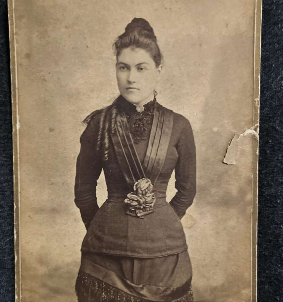 A sepia-toned vintage photograph of a young woman with dark hair styled up, wearing a high-necked, long-sleeved Victorian-era dress with decorative detailing and ribbons, standing against a plain backdrop.