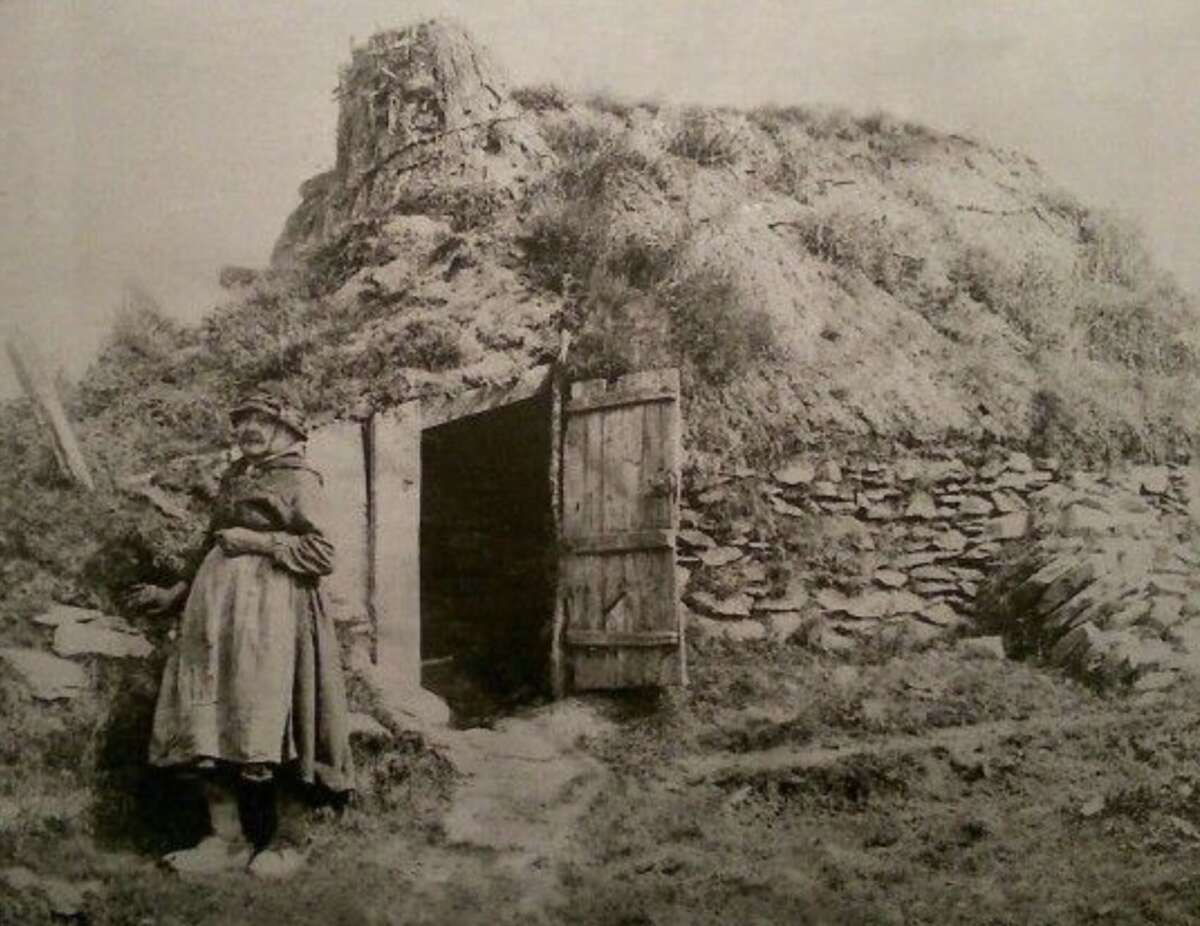 A woman stands outside the entrance of a traditional sod house built into a grassy mound, with a wooden door open behind her and stone walls visible on the side. The image appears old and is in black and white.