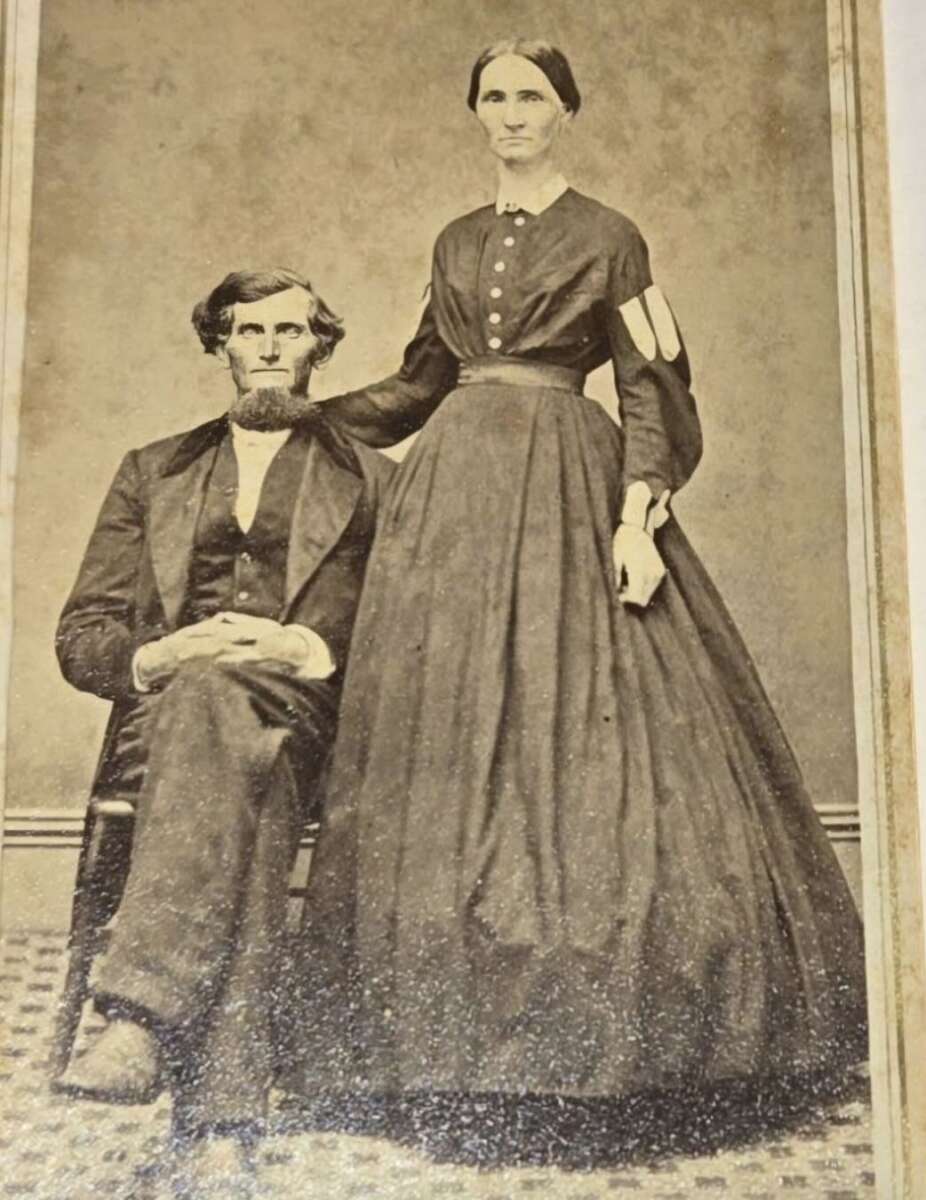 A sepia-toned vintage photograph of a seated man in a suit and a standing woman in a long, dark dress with buttons, resting her hand on his shoulder, both looking at the camera.