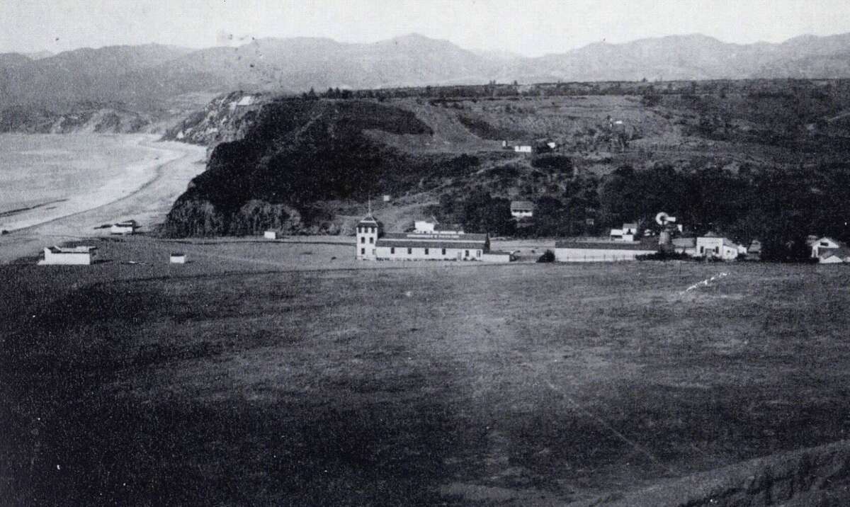 Black and white photo of a coastal landscape with cliffs, a sandy beach, a few buildings including a central long structure, and open grassy fields, with mountains in the background.