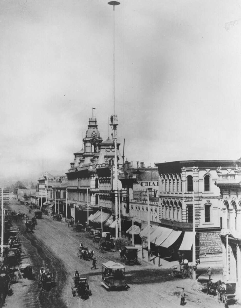 Historic black and white photo of a wide dirt street lined with horse-drawn carriages and old buildings, featuring a tall pole with a round disk at the top, possibly an early streetlight, in a late 1800s town scene.