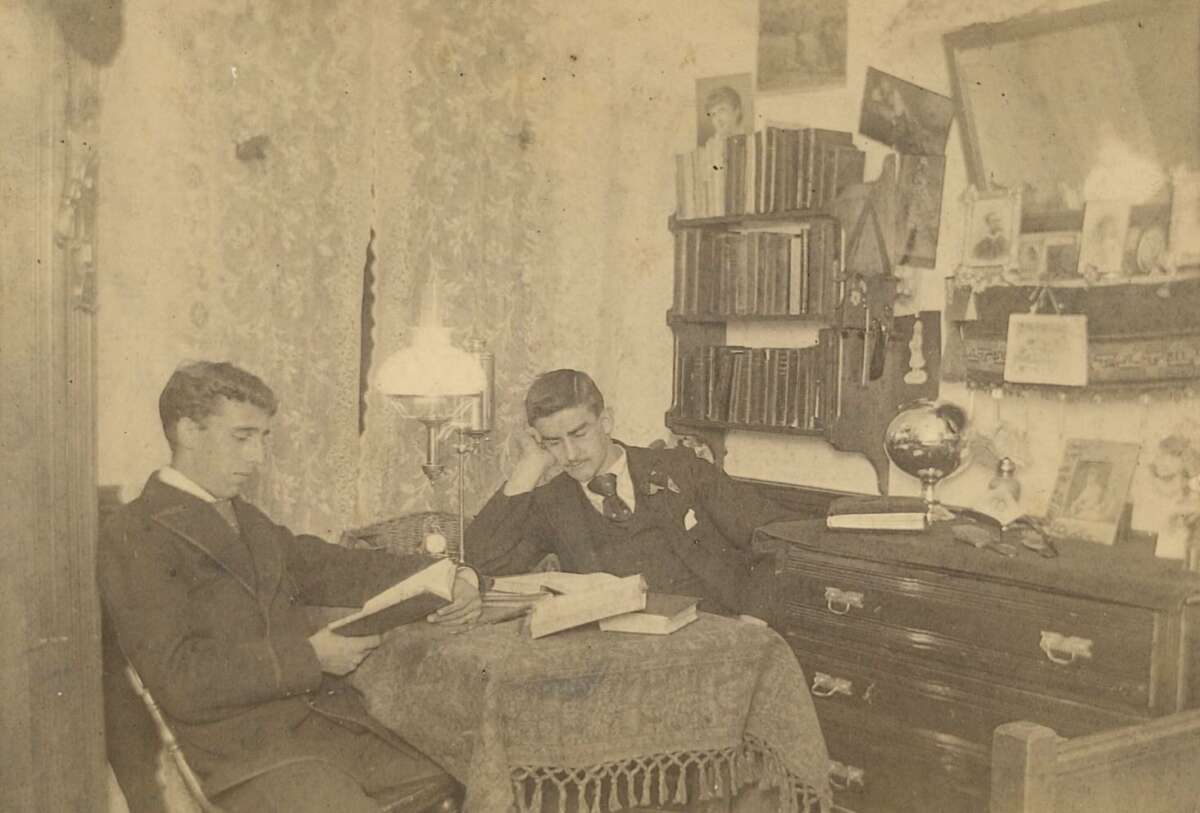Two men in suits sit at a table covered with a fringed cloth, reading books. The room has patterned curtains, shelves filled with books, framed photos, a lamp, and papers on a dresser. The atmosphere is studious and vintage.