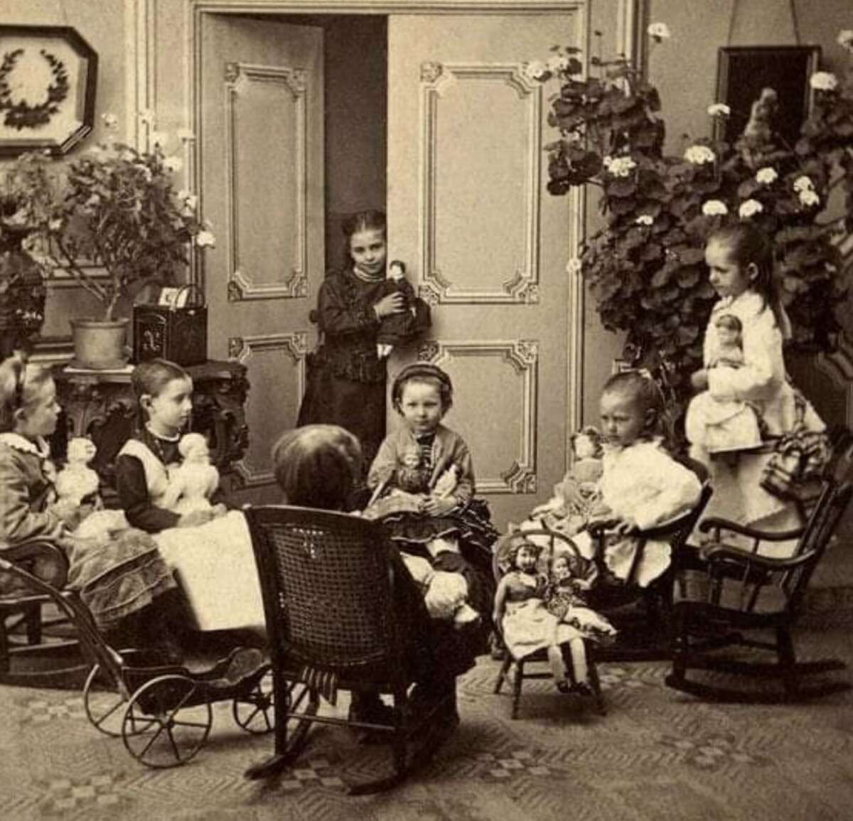 A vintage sepia photo shows seven young girls in dresses gathered indoors, some seated and holding dolls, with one girl standing by a door and plants in the background. The scene has an old-fashioned, Victorian-era atmosphere.