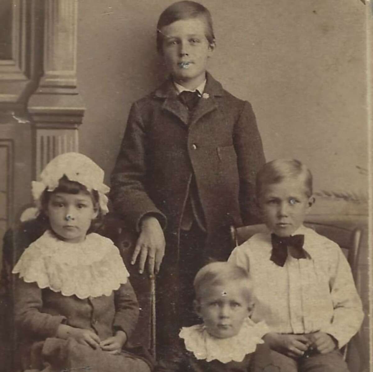 Sepia-toned vintage photograph of four young children in formal clothes; two boys standing and two sitting, one girl wearing a lace bonnet and all dressed in white collars. The background features ornate wooden paneling.