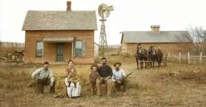 A family of six poses in front of a wooden farmhouse with a windmill in the background. Two horses are hitched to a wagon nearby; the adults and children wear period clothing, suggesting a rural late 19th-century setting.