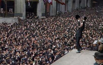 A large crowd of people in early 20th-century clothing gathers in front of a building. A man on a raised platform raises his hat in the air, with many in the crowd waving or cheering.