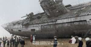 A large, damaged German submarine U-118 is washed ashore on a pebbled beach at Hastings, surrounded by people, including children and adults, who are observing the scene. Text on the image identifies the submarine and location.