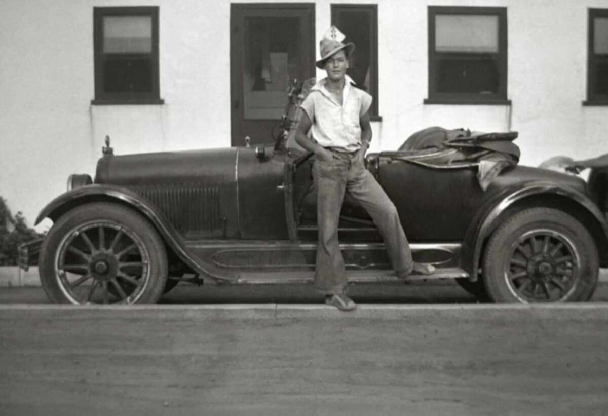 A young man wearing a hat and casual clothes leans against a vintage car parked in front of a white building with two windows.