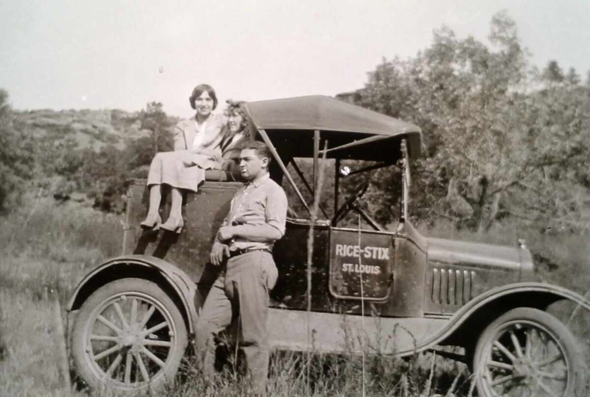 A vintage black-and-white photo shows a woman sitting on the hood of an old car labeled "RICE-STIX St. Louis," with a man standing in front. They are outdoors in a grassy, rural area with trees in the background.