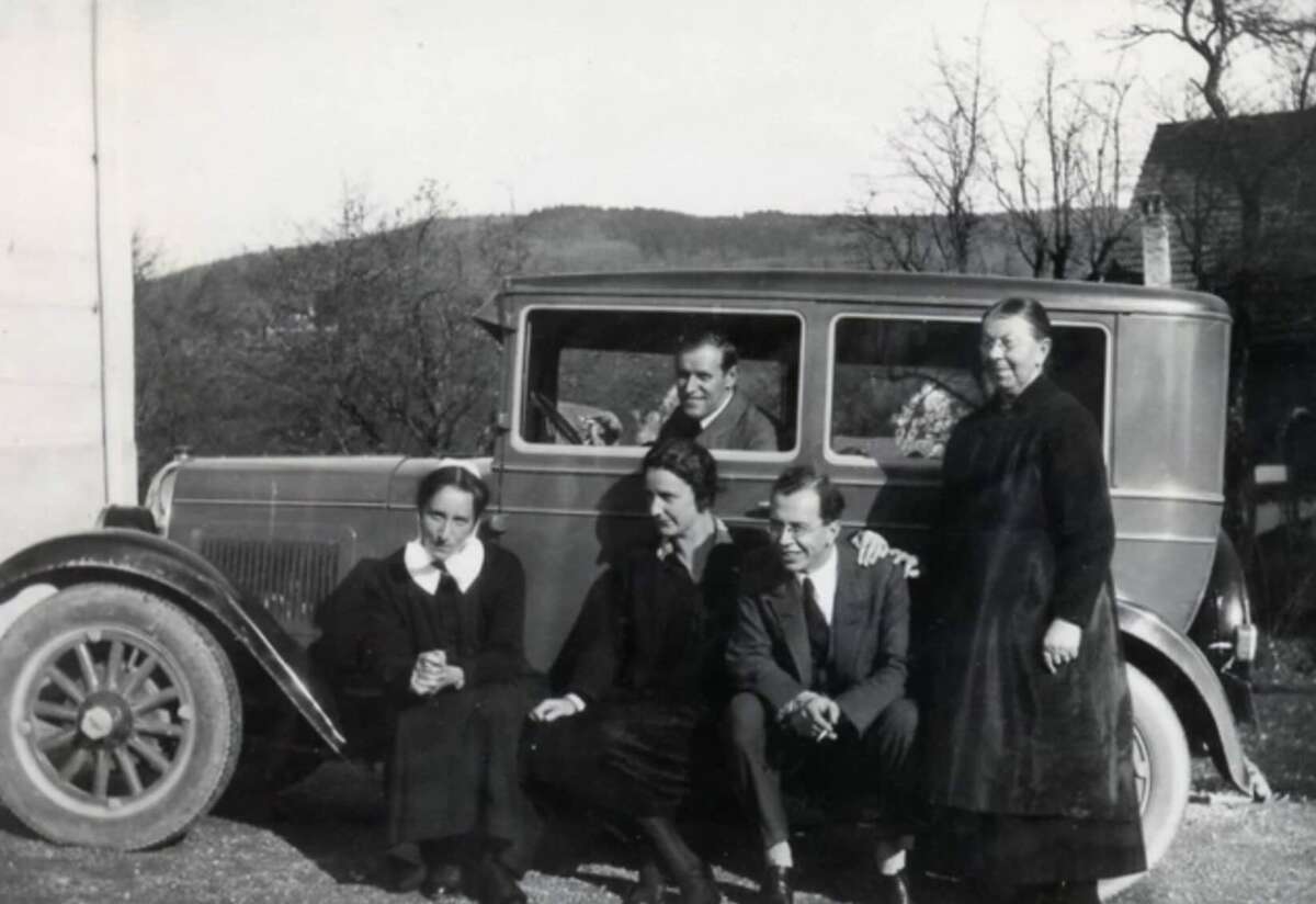 Five adults pose beside a vintage car; four are standing or crouching by the door while one sits inside. Bare trees and hills are visible in the background, suggesting it is autumn or winter. The photo is in black and white.