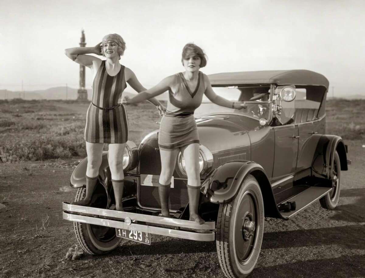 Two women in 1920s swimsuits stand on the front bumper of a vintage car in an open landscape, one shielding her eyes as she looks into the distance, the other holding her arm for balance.