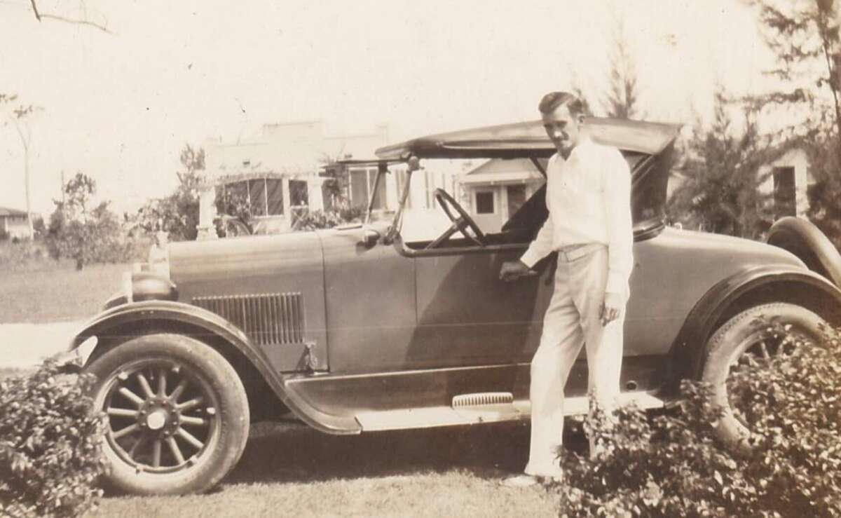 A man in light-colored pants and shirt stands beside a vintage convertible car, smiling at the camera. The scene is outdoors, with houses, trees, and bushes visible in the background. The photo is sepia-toned.