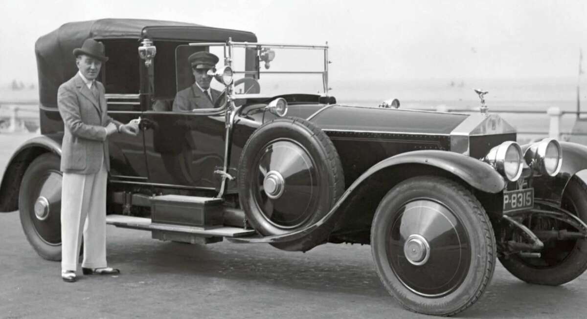 A man in a suit stands beside a vintage luxury car, while a uniformed chauffeur sits behind the wheel. The classic car features large, spoked wheels and a long hood, with a beach or seaside area in the background.