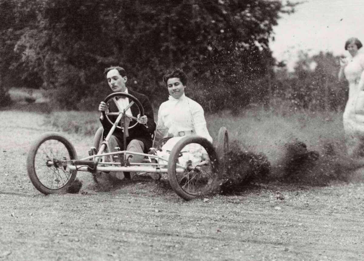 Two people ride in an early open-frame car, kicking up dirt as they speed along a road. The driver wears a suit and bow tie, while the passenger wears a blouse. Trees and a blurred person appear in the background.
