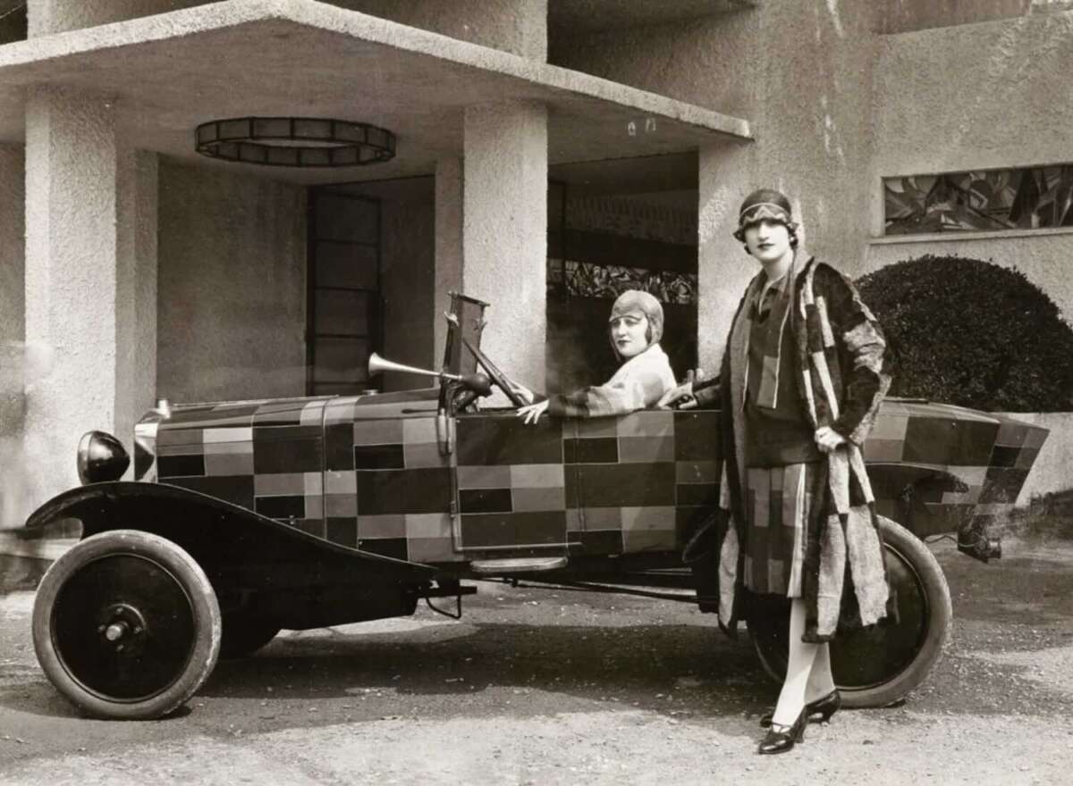 Two women in 1920s attire pose with a vintage car covered in a checkered pattern; one woman is seated in the driver’s seat while the other stands beside the car in front of a modernist building.