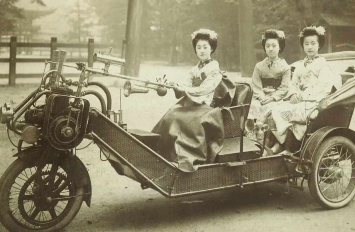 Three women wearing traditional Japanese kimonos sit on a vintage, open three-wheeled vehicle. The driver in front holds a horn, and the two passengers sit behind her. The background is an outdoor park or garden setting.