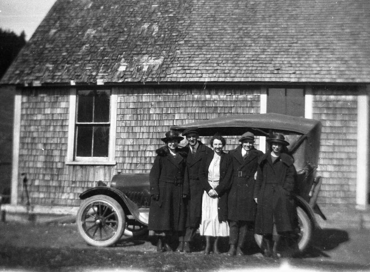 Five people stand in front of an old wooden house and a vintage car. The group is dressed in coats and hats, and the setting appears to be early 20th century. The mood is formal and the photo is black and white.