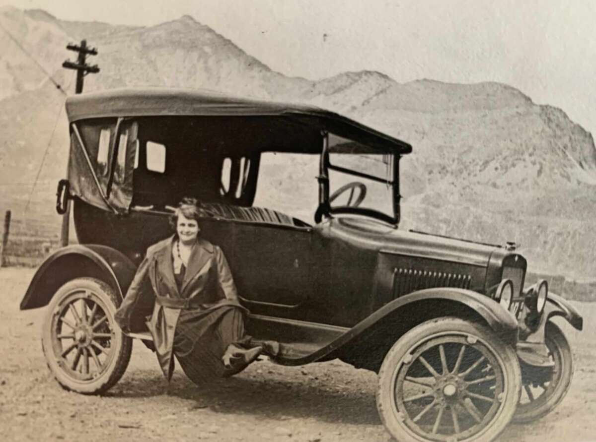 A woman in a coat and dress smiles while sitting on the running board of a vintage car, with mountains and a telephone pole in the background. The photo is in black and white.