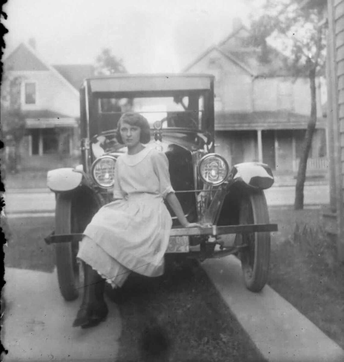 A young woman in a light dress sits on the bumper of an old-fashioned car parked on a driveway, with early 20th-century houses and trees in the background. The photo is black and white.
