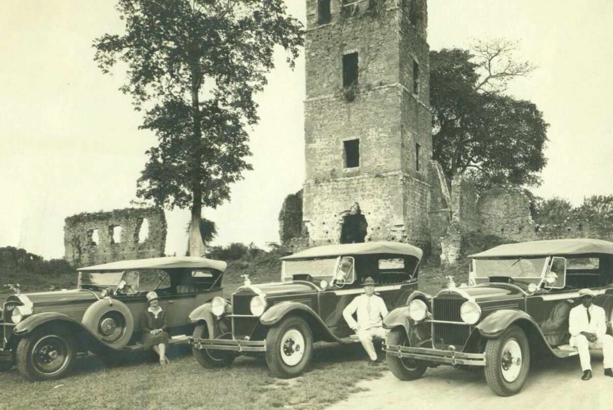 Three vintage cars with their roofs down are parked in front of an old, partially ruined stone tower. Four people in light clothing and hats are sitting or leaning on the cars. Trees and more ruins are visible in the background.