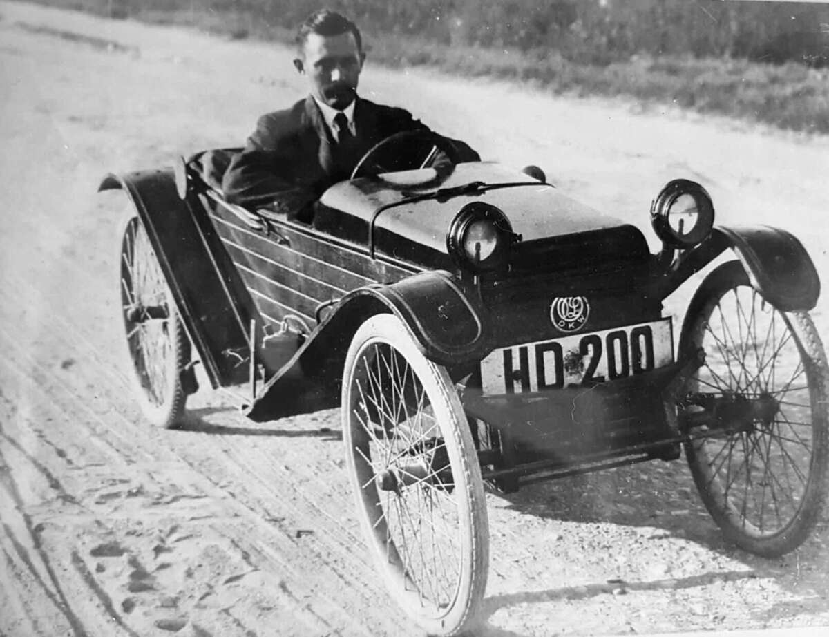 A man in a suit drives an early 20th-century, three-wheeled vintage car on a dirt road. The car has spoked wheels, round headlights, and a license plate reading "HD-2001.