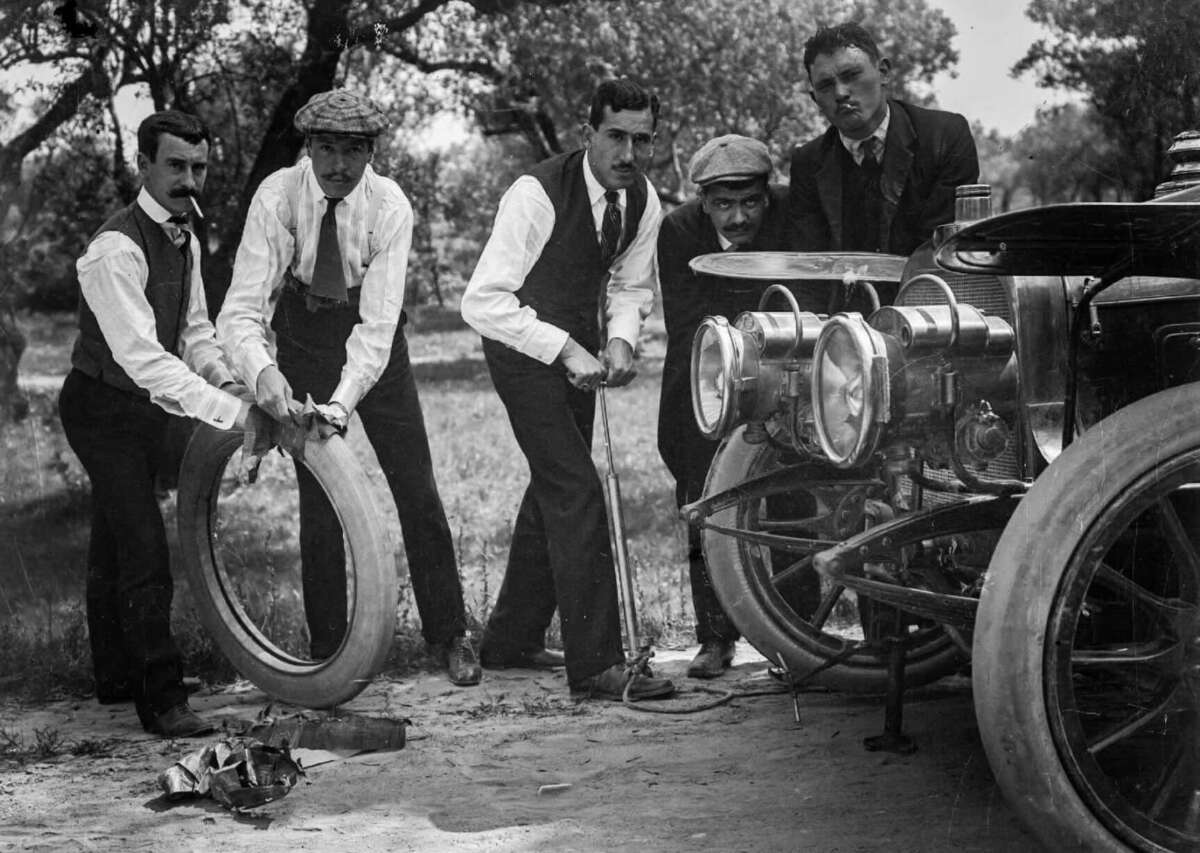 Five men in early 1900s attire work together to change a tire on a vintage car, with one holding the spare tire and others assisting, surrounded by trees and grassy terrain.