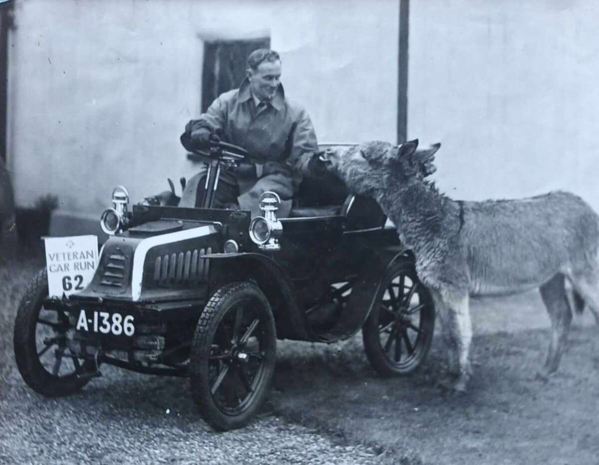 A man sits in a vintage car, smiling and reaching toward a donkey standing next to the vehicle. The car displays the number plate "A-1386" and a sign reading "Veteran Car Run 62.