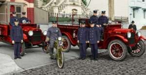 A vintage photo shows uniformed firefighters standing by two red fire trucks and one on a motorcycle on a cobblestone street in front of historic buildings.