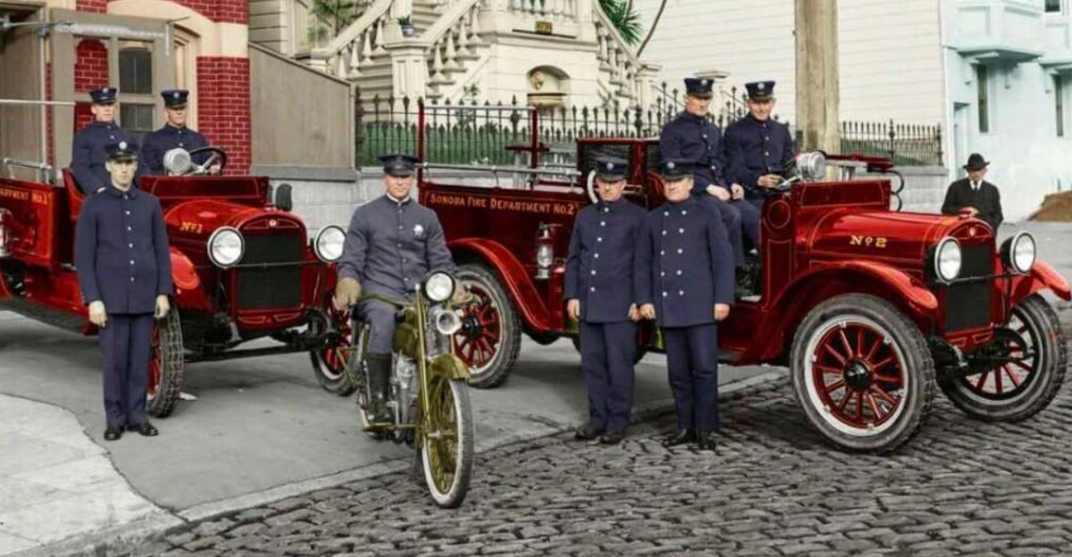 A vintage photo shows uniformed firefighters standing by two red fire trucks and one on a motorcycle on a cobblestone street in front of historic buildings.