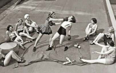 Eight women in vintage swimwear sit on a rooftop, watching two women sparring with boxing gloves in the center. Props like balls and clubs are scattered on the ground near them.