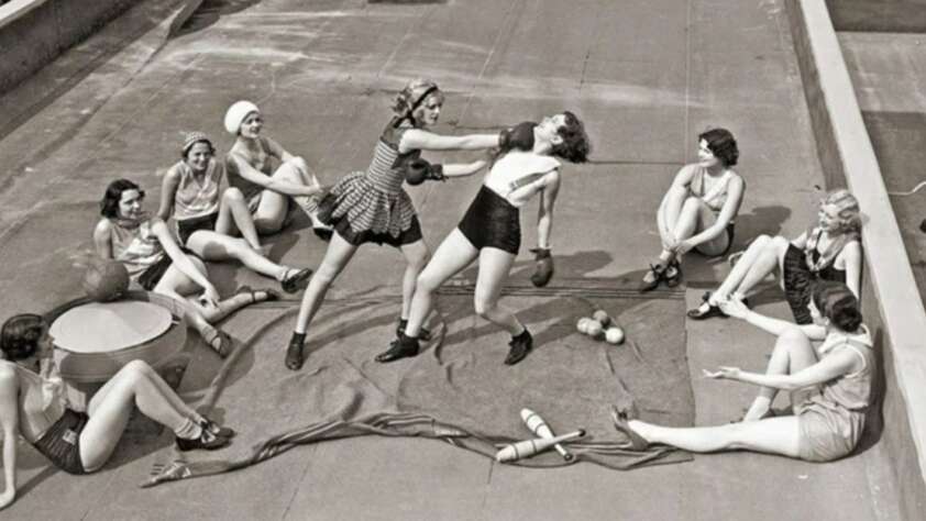 Eight women in vintage swimwear sit on a rooftop, watching two women sparring with boxing gloves in the center. Props like balls and clubs are scattered on the ground near them.