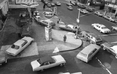 Black and white photo of cars lined up and crowded around a Gulf gas station on a city street corner, likely during a fuel shortage. One person is walking on the sidewalk near the station.