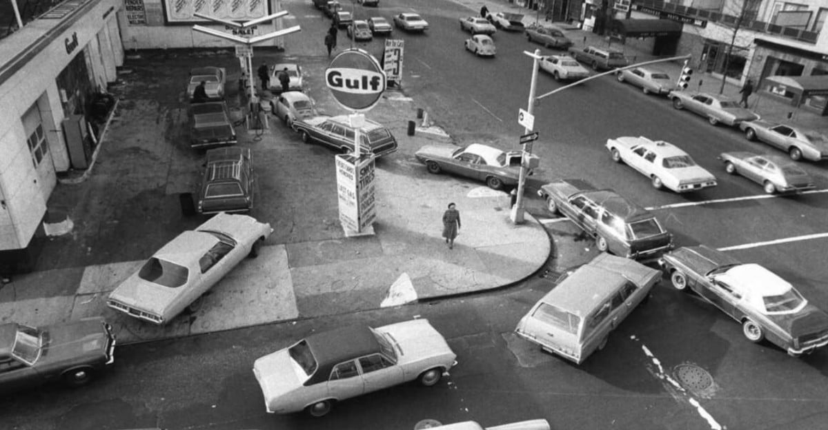 Black and white photo of cars lined up and crowded around a Gulf gas station on a city street corner, likely during a fuel shortage. One person is walking on the sidewalk near the station.