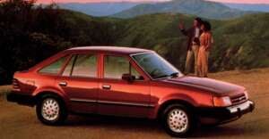 A red 1980s hatchback car is parked on a scenic overlook with mountains in the background, while two people stand nearby, talking and admiring the view at sunset.