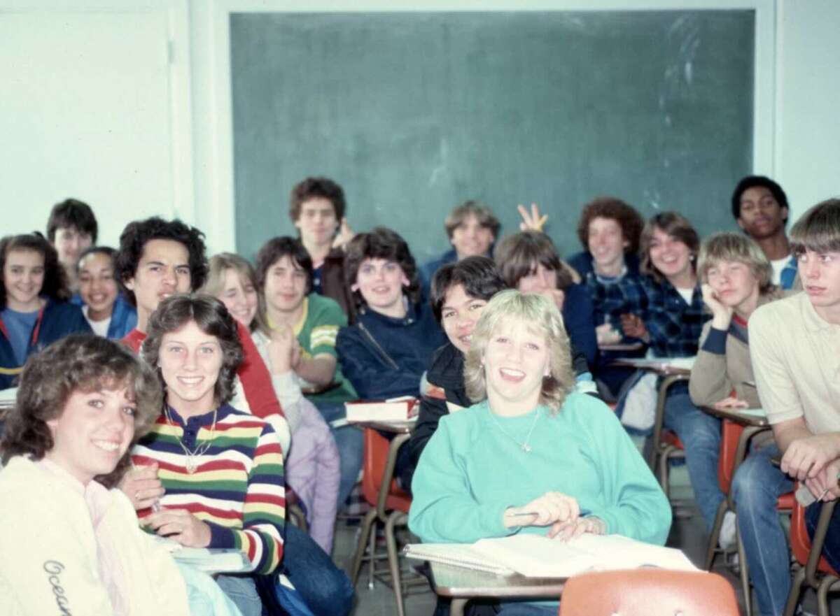 A group of smiling teens sit at desks in a classroom, posing for a photo in front of a chalkboard. Most of them are wearing casual clothes, and some are making peace signs or laughing.