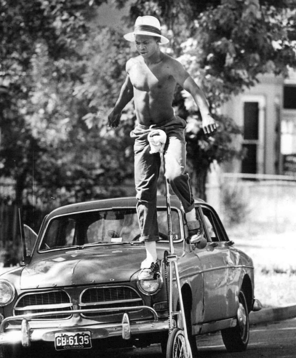 A shirtless man wearing a hat and rolled-up pants balances on top of a vintage car’s hood while stepping onto tall stilts, with trees and houses visible in the background.
