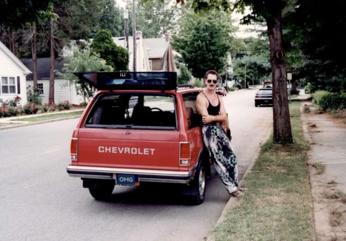 A person in sunglasses and patterned pants leans against a red Chevrolet station wagon parked on a suburban street, with the rear hatch open and trees and houses in the background.
